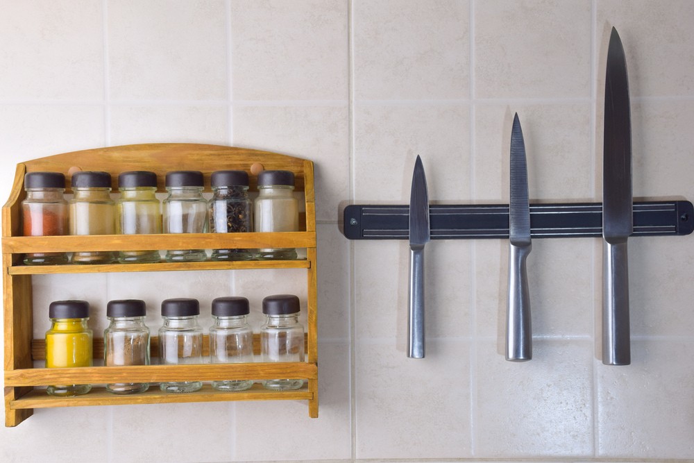 A set of jars with different spices and a set of three steel knives hang on the ceramic wall
